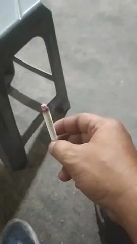 A hand holds a lit cigarette, the tip glowing red. The hand moves slightly, causing the cigarette to flicker. The background is out of focus, but appears to be an indoor setting with a concrete floor and a dark object, possibly a chair, partially visible.