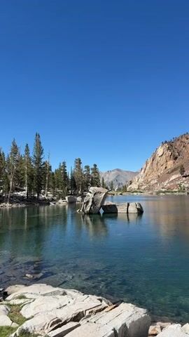 This is a vertical video of a calm lake with clear, turquoise water. Large, gray rocks are visible in the foreground, some partially submerged. In the middle ground, two large rocks emerge from the water. The lake is surrounded by a treeline of tall, green pine trees on the left, and a steep, rocky mountain slope on the right. The sky is a clear, bright blue.
