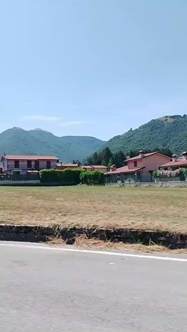 The video shows a side view of a landscape with houses, mountains, and a road. The houses are pink with red roofs and are surrounded by green hedges and trees. There are some dry grass areas in front of the houses. In the background, there are mountains with some vegetation. The sky is clear and blue. A white street light is visible in the middle right of the frame. The camera appears to be moving, likely from a vehicle.