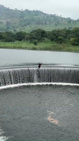 The video shows a person standing on the edge of a circular waterfall, appearing to be preparing to jump. The waterfall is a large, man-made structure with water cascading over its edge into a circular pool below. The surrounding environment is lush and green, with trees and hills visible in the background. The sky is overcast, suggesting cloudy or rainy weather. The person is dark-skinned and wearing dark clothing. The action is frozen at the moment of anticipation before the jump.