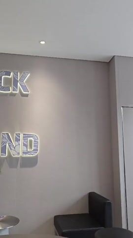 A man is sitting in a chair, looking at his phone. He is wearing a light-colored shirt and dark pants. In front of him is a circular table. Behind the man, on a wall, are large illuminated letters that spell out "LUCK IN HAND." The wall is a light gray color. There are two other tables visible in the foreground, one of which is partially obscured by the frame. The environment appears to be an indoor space, possibly a cafe or waiting area, with overhead lighting.