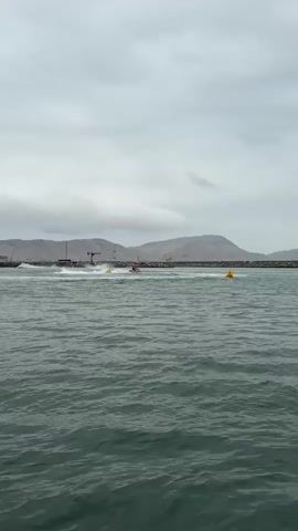 The video shows a view from a body of water, possibly the sea, looking towards a rocky breakwater under a cloudy sky. In the distance, there are hills or mountains. On the breakwater, there are several boats, some of which appear to be in motion, creating splashes in the water. There are also yellow buoys visible in the water near the breakwater. The overall scene suggests a marine environment with some activity taking place.