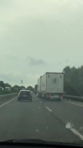 The video shows a view from inside a vehicle traveling on a highway. It is daytime and the sky is overcast. Traffic is visible, including a white semi-trailer truck in the right lane and a dark-colored car in the left lane. There are other vehicles further ahead, suggesting a traffic jam or slow-moving traffic. On the left side of the highway, there is a barrier and trees. A red and white windsock is visible on a pole on the left side of the road. The windshield of the vehicle has some water droplets or smudges on it. The video is shaky, indicating movement.