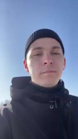 A man with short dark hair and light eyes, wearing a black beanie and a black hooded jacket, is recording a selfie video outdoors on a sunny day. The sky is clear and bright blue.  A snow-covered ground and a paved road are visible.  Some sparse trees are in the background.  He's wearing white earbuds.  He seems to be looking at the camera, moving it slightly to keep his face in the frame. The camera angle is tilted upwards at times, emphasizing the sky.