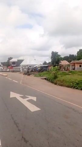 The video shows a roadside scene on a partly cloudy day. A gray asphalt road with white lane markings is visible.  A two-story building, "BARZAQUETA PLAZA", with retail stores is on the right side of the road. Some people are standing and walking around in front of the stores.  There are a few vehicles parked and red canopies set up in front of the building.  To the side of the building, there are some smaller buildings and vegetation. Parts of the building and other objects nearby are covered with white tarps or fabric. The camera is moving along the road and past the building.