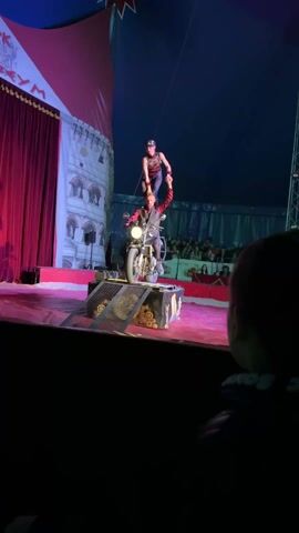 A medium shot shows a circus performance on a stage. A man in black and red clothes sits on a motorcycle, which is positioned on a decorated platform at the center of the stage. A woman in similar attire is standing on his shoulders, striking a pose. In the foreground to the right is the blurry silhouette of a person, presumably an audience member. In the background, behind the performers, is a drawn backdrop that appears to be of a building. Red curtains hang to the left of the backdrop. An audience is seated in the background. The lighting is dramatic, with the stage illuminated in bright pink and blue hues, emphasizing the action while the surrounding area remains relatively dark.