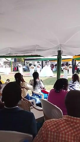 The video captures a wedding scene from under a large white tent, where guests are seated, facing a couple dancing on a green lawn. The bride is wearing a large white dress. The guests are sitting in white chairs, some are wearing casual clothing, and one woman is using a phone to take photos. The scene is decorated with flower arrangements.