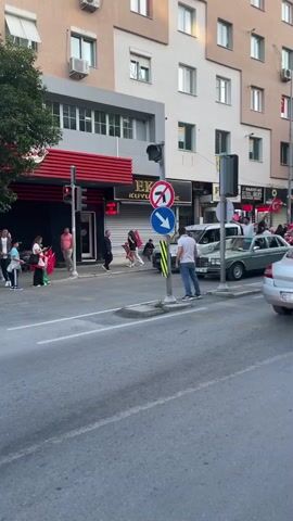 The video shows a street scene in what appears to be a Turkish city. Several people are standing on the sidewalks, some waving Turkish flags. There is moderate traffic on the street, with cars passing by, including a white Renault Clio visible in the foreground. The buildings along the street are multi-story and have a mix of residential and commercial uses. Shop signs are visible in Turkish. The sky is clear and the lighting suggests it is late afternoon. There is a traffic signal with road signs next to it.