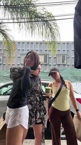 Four young people are standing outdoors. From left to right, there are three young women and one young man. The first woman is wearing a black long-sleeve shirt and white shorts. The second woman is wearing a black and white floral dress and is holding a drink in her hands. The third woman is wearing a yellow tank top and brown pants and is carrying a brown paper bag. The young man is wearing a black t-shirt, shorts, and a baseball cap. Behind them is a silver car parked in front of a gray metal building with windows. A palm tree is visible in the upper left corner of the frame. The young man walks into the shot from the right and they all move slightly in place, posing and smiling.