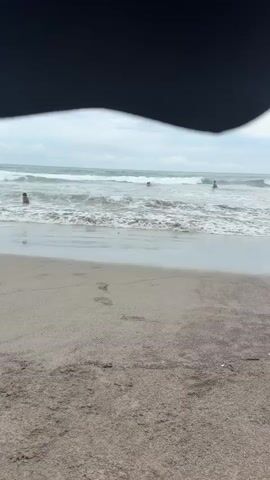 A short video clip shows a sandy beach with gentle waves rolling onto the shore. The sky is overcast. Several people are visible in the water, swimming or playing in the waves. The foreground of the video shows a patch of wet sand with footprints. A dark, curved object, possibly a shadow or part of an umbrella, partially obscures the top of the frame.