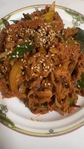 A close-up shot focuses on a white plate filled with what appears to be Korean spicy pork stir-fry, or Jeyuk Bokkeum. The meat is thinly sliced and coated in a reddish-brown sauce, mixed with visible pieces of green onion and onion. The dish is generously sprinkled with white sesame seeds. The plate has a decorative green and tan border. The background is slightly out of focus, showing a portion of a white table and a patterned tray with blue leaf designs. The lighting is bright, suggesting an indoor setting, possibly a kitchen or dining area. The camera slowly pans and zooms in slightly, offering a detailed view of the food.
