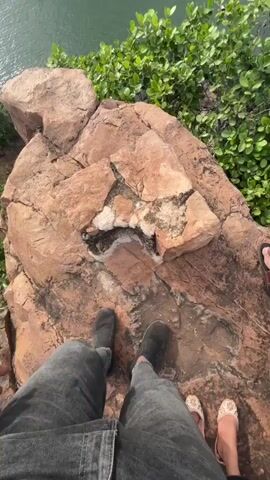 The video begins with a first-person perspective looking down at a person's feet wearing dark boots and grey jeans standing on a rocky cliff edge. Green foliage is visible on the right. The camera then pans out to reveal a wide shot of a body of water, possibly a river or lake, with two waterfalls cascading down a cliff face in the distance. The surrounding landscape is lush and green, with rocky outcrops and trees. The sky is cloudy. The shot focuses on the natural scenery.