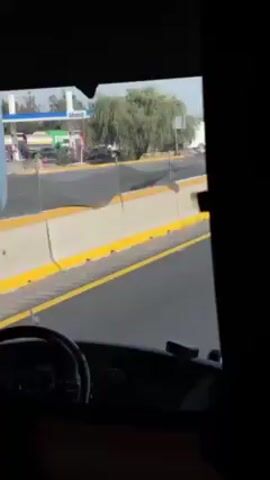 The video shows a view from inside a vehicle, likely a truck or bus, driving on a highway. The road is divided by a concrete barrier with a yellow stripe.  Outside the vehicle on the other side of the barrier, a Mobil gas station and other vehicles, including semi-trucks, can be seen. The sky is overcast. The vehicle's dashboard and steering wheel are visible in the foreground.