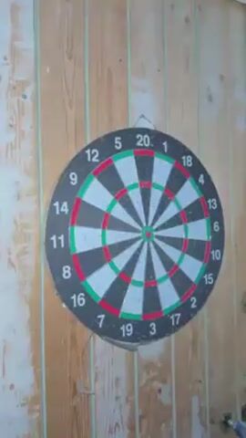 The video shows a dartboard mounted on a wooden wall. Four darts are thrown at the dartboard. One dart lands in the "18" section, one lands near the "14" section, one lands near the "5" section, and one dart falls on the floor.