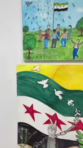 The video shows a selection of artworks hanging on a white wall. The artworks appear to be paintings and drawings made by children, with themes related to Syria. Some artworks depict scenes of destruction and conflict, while others express hope for peace and unity. One painting shows children holding hands in a circle under the Syrian flag, with roses falling from the sky. Another painting depicts a chained hand giving a peace sign in front of a Syrian flag. One shows a boy sleeping within the borders of Syria, which is cracking. The setting suggests an art exhibition or display showcasing children's perspectives on the Syrian conflict.