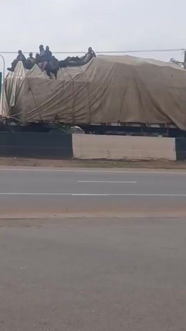 The video shows a large truck, partially covered by a tan tarp, driving on a multi-lane highway. Several people are sitting on top of the covered cargo area. The truck is green and the road is paved. The sky is overcast.