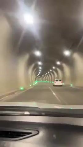 A vehicle's dashboard is visible, with a view of a brightly lit tunnel through the windshield. Another car is visible ahead in the same lane. The tunnel is illuminated by overhead lights and a series of green lights along the road's edge. The road is marked with yellow lane lines. The tunnel walls are a light color, possibly concrete.