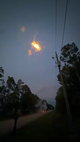 The video captures a nighttime scene of fireworks being set off. The sky is dark blue, and in the upper part of the frame, a large burst of red and yellow fireworks illuminates the sky. Below the fireworks, silhouetted trees line the sides of a dark road. In the foreground, on the right side of the frame, a utility pole with wires is visible. On the road, several small figures, presumably people, are present, and smoke from previously ignited fireworks drifts upwards. The overall atmosphere suggests a festive but somewhat hazy environment due to the fireworks.