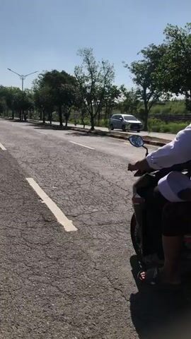 The video shows a view from a moving vehicle on a cracked asphalt road. Two motorbikes are visible, each carrying two people. The first motorbike, on the left, has a rider with a red helmet and a passenger wearing a white shirt. The second motorbike is further down the road, also with two occupants. Trees line the right side of the road, beyond which is a fence and more vegetation. A car is seen in the distance on what appears to be another road. The sky is clear and blue, indicating a sunny day.