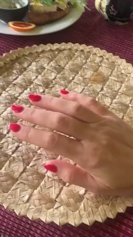 The video shows a close-up shot of a person's hand with bright red nail polish resting on a woven placemat. The placemat is round and made of natural fibers, with a geometric pattern. In the blurred background, a white plate with food, a slice of orange, and a cup can be seen on a dark red tablecloth. The hand moves slightly, showing the fingers and palm.