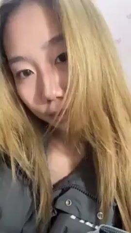 A person with long, light-brown hair, wearing a gray jacket, looks at the camera. They place a hand on their cheek and then move their hair. The background is a white wall with a light pink pattern.