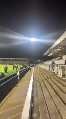 The video shows a view of a soccer stadium at night. The field is brightly lit by floodlights. There are a few people in dark clothing on the field, walking around. The stands are mostly empty, with a few people scattered here and there. The perspective is from the edge of the field, looking across to the stands on the other side. The video is steady, and the focus is clear.