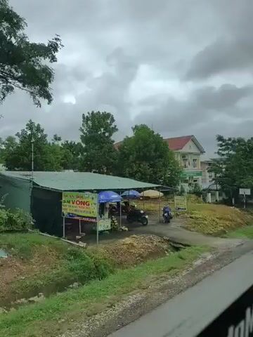 The video shows a view from a moving vehicle traveling along a road. The scene is predominantly outdoors with overcast skies and lush green trees. On the left side of the road, there is a building with a white facade and a red roof, possibly a residential building or an office. In front of it, a white SUV is parked. Further down the road, another building with a similar architectural style is visible. There are also several parked motorcycles under a sheltered area. The ground appears wet, suggesting recent rain. The overall environment seems to be a semi-rural or suburban area. There are no people clearly visible in focus or interacting within the frame, except for the distant figures under the shelter. The focus is on the landscape and the buildings along the route.