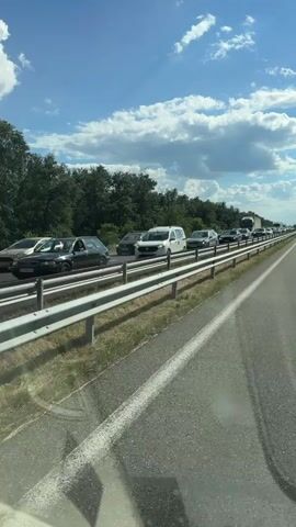 The media file shows a traffic jam on a highway, likely in Bozduganovo, Bulgaria. The scene is set outdoors on a sunny day with scattered clouds. The highway is multi-lane, with a metal guardrail separating the lanes of traffic from the grassy shoulder and trees lining the distant background.

The primary objects are cars and trucks, all appearing to be stationary or moving very slowly. Several passenger cars of various makes and models are visible, along with at least one large semi-truck. The vehicles are lined up bumper-to-bumper, indicating a significant traffic delay.

The time of day appears to be daytime, judging by the bright sunlight and the blue sky. The weather seems pleasant, with the sun shining and a gentle breeze suggested by the clouds. There are no people clearly visible in the foreground or immediately around the cars. The overall mood conveyed is one of frustration or impatience due to the traffic standstill.

There is some visible text on the side of the semi-truck: "ALP ÖZER". This suggests the truck belongs to a logistics or transportation company. The image is taken from inside another vehicle, likely at the same level as the traffic, looking forward along the highway.