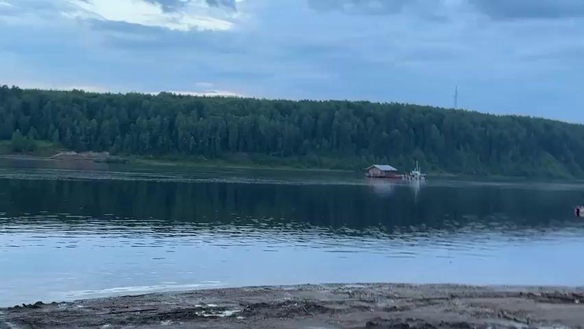 The video opens with a wide shot of a serene river under a cloudy sky. A lush, green forest covers the distant bank, reflecting perfectly in the calm water. In the middle of the river, a small, flat-bottomed barge with a wooden structure on top is visible, accompanied by a smaller white boat. As the camera pans to the right, the shoreline comes into view, revealing a muddy bank. Further along the bank, a ferry is docked. The ferry is a utilitarian vessel with a large flat deck for vehicles and a small cabin structure at the rear. The ramp of the ferry is lowered to the ground, suggesting it is either preparing for departure or has just arrived. The overall atmosphere is peaceful and tranquil.