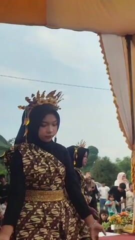 A vertical medium shot shows a young woman in traditional attire dancing on a stage outdoors. She wears a black hijab, a brown and white batik-patterned dress with a gold belt, and a golden headdress with dangling ornaments. Her hands are held up and she moves them gracefully. Another dancer in similar attire is visible behind her. The background shows a crowd of spectators and a tent with yellow and white drapes. The sky is cloudy.