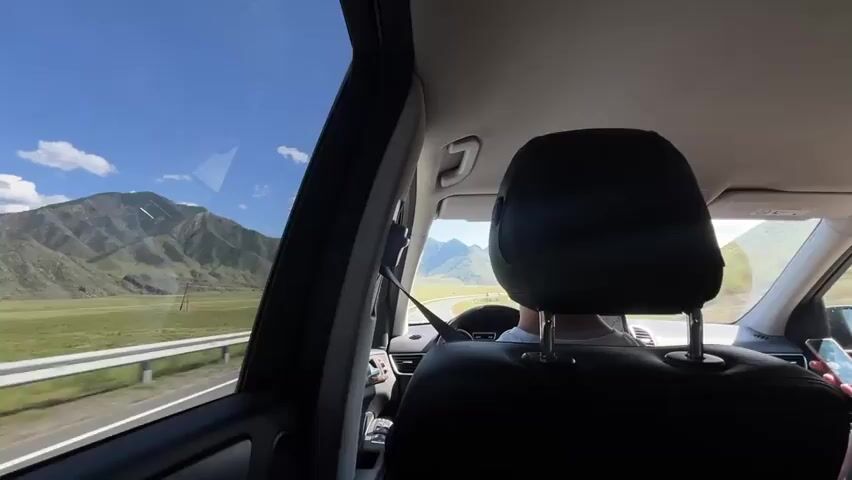 The video shows the interior of a car from the backseat, looking forward. The camera is positioned behind the headrests of the front seats. A person's head and shoulders are visible in the foreground, with the back of their head and neck in focus. This person appears to be wearing a white t-shirt. They are sitting in the driver's seat, with their hands on the steering wheel. The steering wheel is visible, along with part of the dashboard. Outside the car, through the windshield and side windows, a mountainous landscape is visible under a bright blue sky with some clouds. The car is driving on a road with a white guardrail to the left. The overall impression is a scenic drive through a mountain region.