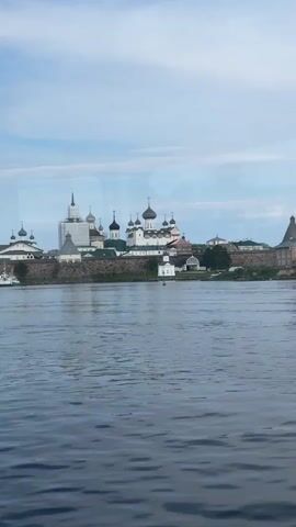 A wide shot shows a body of water in the foreground and a stone fortress with multiple domed buildings in the background. The sky is blue with white clouds. The fortress appears to be a monastery with white walls and a variety of architectural styles. The water is dark blue and has small waves. There is a white boat to the left of the fortress. The overall scene is calm and serene.
