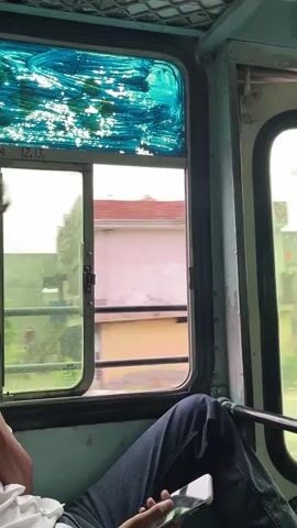 The video is an interior shot from inside a moving vehicle, likely a bus, looking out a window. A person's legs, clad in blue jeans, are visible in the lower right corner of the frame, with their hands holding a smartphone. The window has a sliding pane and is decorated at the top with streaks of blue and white paint or film. Outside the window, buildings and trees are blurred due to the motion, indicating the vehicle is in transit. The scenery suggests a rural or suburban environment.