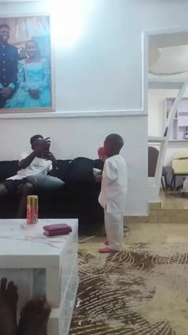 Here's a description of the video:

The video shows a living room setting. A young boy in a white traditional outfit stands in the middle of the room. He is facing a man who is sitting on a black sofa. The man is wearing a white T-shirt and shorts. He's holding a smartphone and seems to be interacting with the child.  A can of drink and a wallet are on a white coffee table in front of the sofa.  On the wall behind the sofa is a framed picture of a couple. The floor is covered with a patterned rug. In the lower left corner of the frame, the viewer can see a pair of bare feet. The lighting in the room is bright, coming from an overhead light.  Behind the sofa, a small section of what appears to be another room is visible.