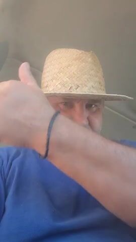 A man wearing a straw hat and a blue t-shirt gives a thumbs up. He is inside a vehicle.