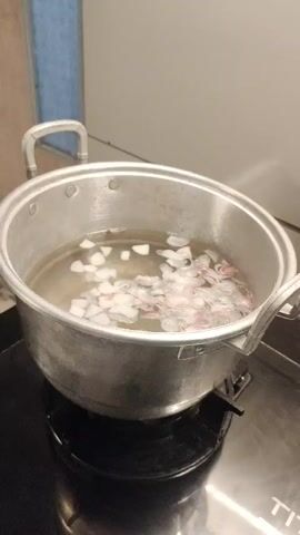 A still shot shows a pot of water with flower petals in it, sitting on a stovetop. The pot is silver and has two handles. The water is clear and some of the petals are floating on the surface, while others are submerged. The stovetop is black and appears to be electric. The background is blurred, but it looks like a kitchen.
