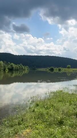A serene landscape video features a calm body of water reflecting a cloudy sky and a tree-lined hill. In the foreground, green grass grows along the water's edge. As the camera pans, a person's back is visible, wearing a white shirt and a light blue cap. They appear to be looking down at the water. The video continues to show the natural scenery, with a rocky cliff face covered in trees and more dense forest on the hillsides. The water remains tranquil, with reflections of the sky and surrounding vegetation. The video concludes with a view of the water and the forested hills.