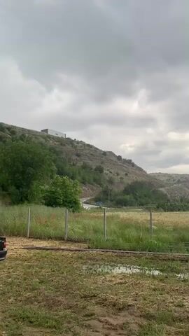 The video captures a static view of a rural landscape in Belo Pole, Bulgaria, during a rainy day. The sky is heavily overcast with dark grey clouds, and the sound of falling rain is prominent throughout the clip.

In the immediate foreground, the ground is muddy with several puddles, suggesting recent or ongoing heavy rainfall. A wire fence with wooden posts stretches across the middle of the frame, separating the muddy foreground from a larger field. This field contains a mix of green and golden vegetation, possibly crops like wheat or barley, and is dotted with patches of bright red poppies.

Behind the field, a winding road is visible, on which a white vehicle and possibly another car can be seen driving. Further in the background, a series of hills rise, covered with sparse green foliage and some trees. On the crest of the left-most hill, a distinctive white building with a flat roof stands out against the dark sky. The overall impression is one of a quiet, somewhat moody landscape under a heavy rain shower.