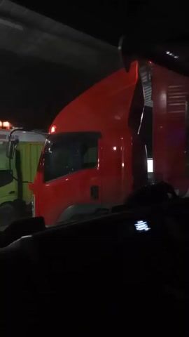 A person lies down, apparently to rest, in the driver's seat of a dark vehicle, placing their bare foot on the side pillar of the windshield. Through the windshield is seen another red truck, parked with its light on. A person in a yellow shirt walks in the background. The area appears to be a covered or indoor space.