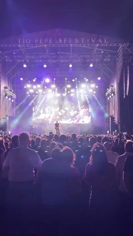 A concert is taking place on a brightly lit stage. The stage has a large screen displaying visuals of performers and the crowd. A band is performing, with a lead singer at the front of the stage. The audience is visible in the foreground, with many people holding up their phones to record the performance. The lighting is predominantly purple and white, with smoke effects adding to the atmosphere. The text "TIO PEPE FESTIVAL" is visible on the stage structure.