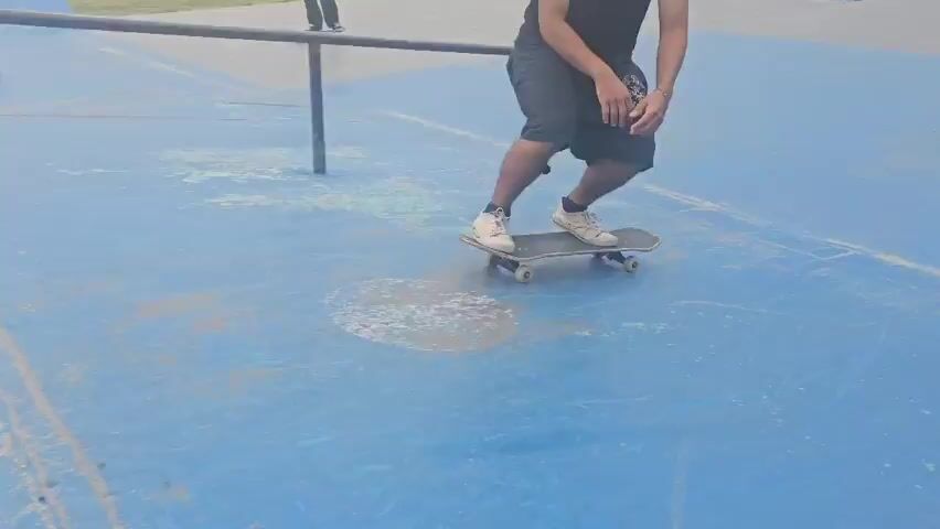 The video captures a person skateboarding in a skatepark. The individual, wearing a black t-shirt, dark shorts, and white sneakers, is seen performing an ollie. The skateboard is a standard model, and the park features a blue surface with various obstacles like metal rails and ramps. Another person can be seen in the background. The lighting suggests it's daytime.