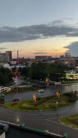 A wide shot captures a city intersection at dusk with a roundabout in the foreground. Cars and buses navigate the wet road, reflecting the streetlights. In the center of the roundabout, a landscaped island features two large, illuminated star-shaped decorations. Beyond the intersection, trees and buildings form a cityscape, with a tall, red-and-white striped chimney visible to the left. The sky is a gradient of orange and gray clouds, suggesting the end of a day with lingering light. The overall scene depicts a moderately busy urban environment transitioning into the evening.