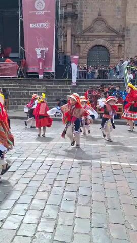 A group of young children dressed in traditional colorful outfits with hats are dancing in a cobblestone square in front of a stone building. They are performing some kind of cultural dance in front of an audience. The children are wearing red, orange, and white clothing with intricate patterns. Some of them have red tassels attached to their outfits. The background shows a stage with banners that have logos and text, a crowd of spectators, and the facade of an old building. The lighting suggests it is daytime.