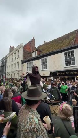A street parade or procession is underway in what appears to be a town or village setting. The scene is crowded, with many onlookers watching and recording the event with their phones. A large, imposing figure dressed in a brown hooded robe and white sleeves stands above the crowd. The figure appears to be on a wheeled platform or cart, as it's being moved along the street. The crowd consists of people of varying ages. The environment features buildings with traditional architecture, and shops are visible. The overall atmosphere suggests a festive and celebratory event.
