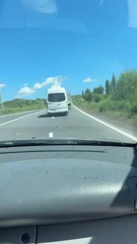 The video shows a first-person perspective from inside a vehicle traveling on a highway. The view is through the windshield, which is cracked. In the distance, a white camper van is driving on the highway. The landscape on either side of the road is green and hilly, with trees and some scattered clouds in the bright blue sky. The camera angle shifts slightly throughout the video, tilting up and to the side, revealing more of the sky and a larger mountain in the background. A traffic sign is visible on the side of the road in the latter part of the video. The sun appears to be shining, indicating it is daytime.