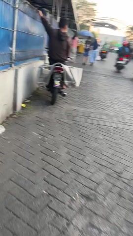 A person is seated on a motorcycle, facing away from the camera. They are wearing a dark hooded sweatshirt. The motorcycle has a large, makeshift cargo carrier attached to the rear. The person is reaching up and to their left, seemingly interacting with something above and to the side of the motorcycle. The motorcycle is parked on a paved walkway. In the background, there is a blue fence or wall, and beyond that, several other people are visible, along with market stalls or vendor carts. The lighting suggests it is daytime, possibly late afternoon or early morning.
