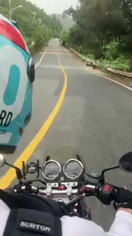 The video shows a first-person view of someone riding a motorcycle on a wet road near the ocean. The rider is wearing a blue helmet, a white shirt, and a black Burton backpack. The motorcycle has two round gauges and is moving along a road with a solid yellow line. The road curves, and on the right side, there is a view of the ocean. Several parked scooters and trees line the roadside. Red and white striped guard rails appear along the road's edge. The sky is overcast. Warning signs are visible near a bend in the road.