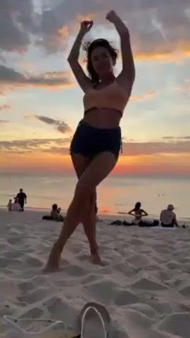 A woman is standing on a sandy beach at sunset. She's wearing a light orange tank top and dark jean shorts. Her dark hair is long and wavy. She turns around, posing in front of the sunset. The sky is cloudy with orange and yellow hues from the setting sun, which is just above the horizon. In the background, there are other people on the beach, some sitting, some standing, and some near the water. In the foreground, a pair of white flip-flops lies on the sand.