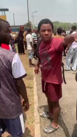 The video shows a crowd of young men on a paved area near some grassy patches and a drainage ditch.  One man wears a maroon t-shirt and shorts and speaks emphatically while gesturing. Another man in a gray tank top is looking at him. Several others are standing nearby, and the crowd extends into the background. A dark-colored car with its driver-side door open is parked halfway down the side of a short grassy bank into the drainage ditch. More of the crowd surrounds it. The overall impression is of a gathering, perhaps an incident has occurred with the car.