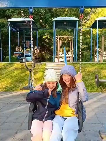 Two young girls are on a swing set. The girl on the left is wearing a light green hat and a dark blue jacket with pink pants. The girl on the right is wearing a blue hooded jacket and light blue pants, with an orange shirt underneath. They are both smiling and laughing as they swing back and forth. The background shows a playground with trees and other playground equipment. The sun is shining and the lighting suggests it is daytime. The camera appears to be positioned in front of and slightly below the girls, looking up at them.